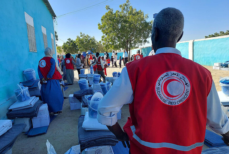 Sudan_Hilfsgueter_fuer_White_Nile  Eine Gruppe von Mitarbeitern des Sudanesischen Roten Halbmonds, die rote Westen mit dem Logo der Organisation tragen, verteilt Hilfsgüter wie Decken, Eimer und verpackte Waren an eine Menschenmenge vor einem leuchtend blauen Gebäude. Die Szene findet unter klarem Himmel statt, mit ordentlich gestapelten Hilfsgütern. Im Vordergrund steht ein SRCS-Mitarbeiter mit dem Rücken zur Kamera.