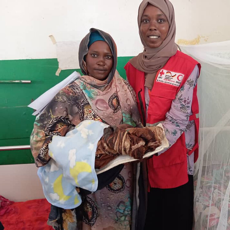 Sudan_Mutter_und_SRCS-Mitarbeiterin Eine Frau in farbenfroher Kleidung hält ein kleines Kind in einer Decke, während sie neben einer Mitarbeiterin des Sudanesischen Roten Halbmonds steht, die eine rote Weste trägt. Beide Frauen lächeln leicht in die Kamera. Im Hintergrund ist ein weißes Moskitonetz über einem Bett sichtbar.