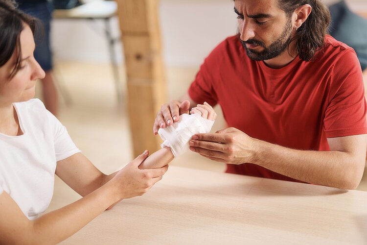 Erste Hilfe Situation: Schnittwunde Erste Hilfe Situation: Versorgung einer Schnittwunde an der Hand, die sich eine Frau beim Gemüseputzen in der Küche zugezogen hat. Der Mann legt eine Mullbinde an.