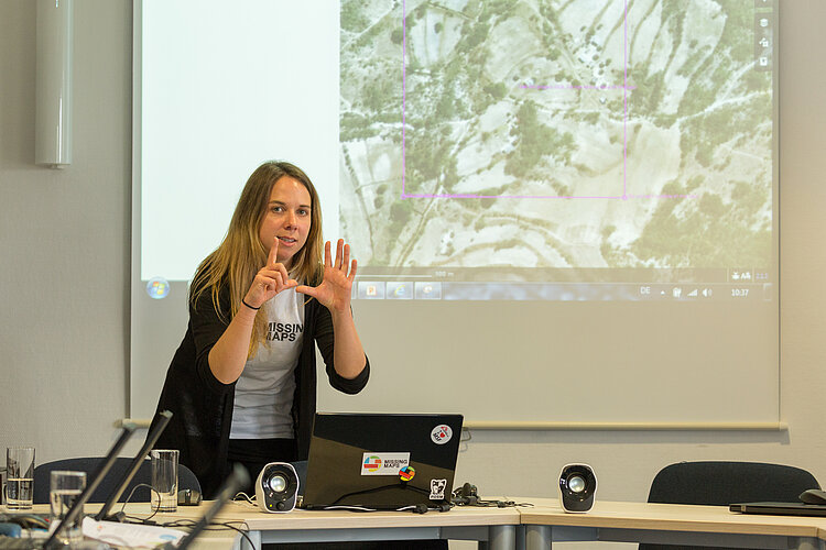 Missing Maps Eine Frau steht vor einer Projektionsfläche und hält eine Präsentation. Sie trägt ein T-Shirt mit der Aufschrift "MISSING MAPS" und eine schwarze Strickjacke. Auf dem Projektionsbild ist eine Landkarte zu sehen. Vor ihr steht ein Laptop, auf dem ebenfalls Aufkleber von "MISSING MAPS".