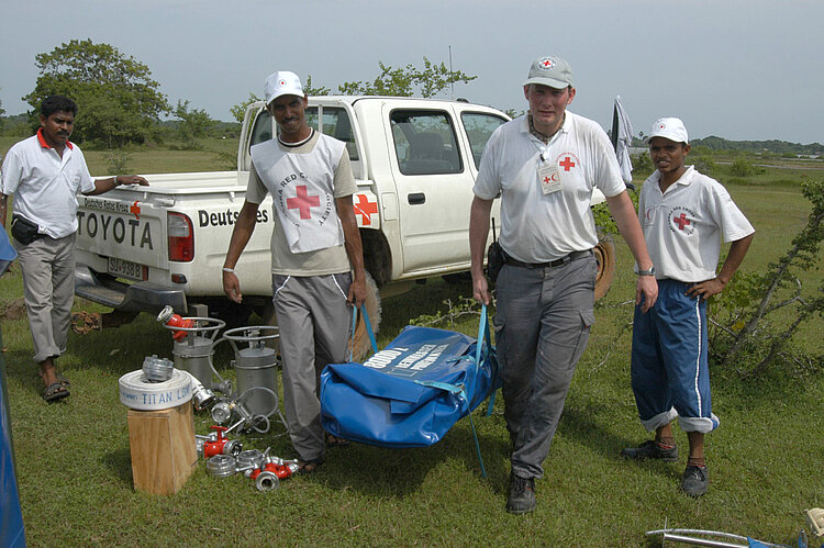 20 Jahre Tsunami - Helfer Sri Lanka Vier Mitglieder des Roten Kreuzes stehen neben einem Hilfsfahrzeug mit Wasseraufbereitungsgeräten in Pottuvil, Sri Lanka. Zwei von ihnen tragen eine blaue Tasche mit Equipment, während verschiedene Ausrüstungsgegenstände auf dem Boden verteilt sind.