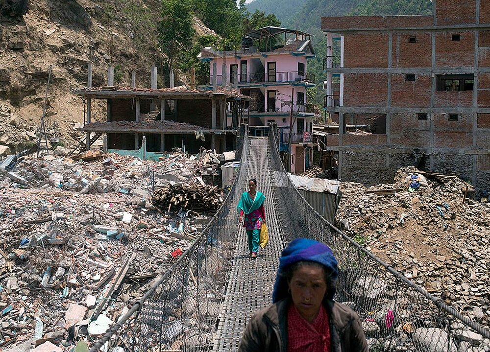 Menschen gehen über eine Hängebrücke, im Hintergrund die zerstörete Ortschaft Singati in Nepal