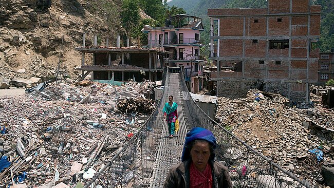 Menschen gehen über eine Hängebrücke, im Hintergrund die zerstörete Ortschaft Singati in Nepal