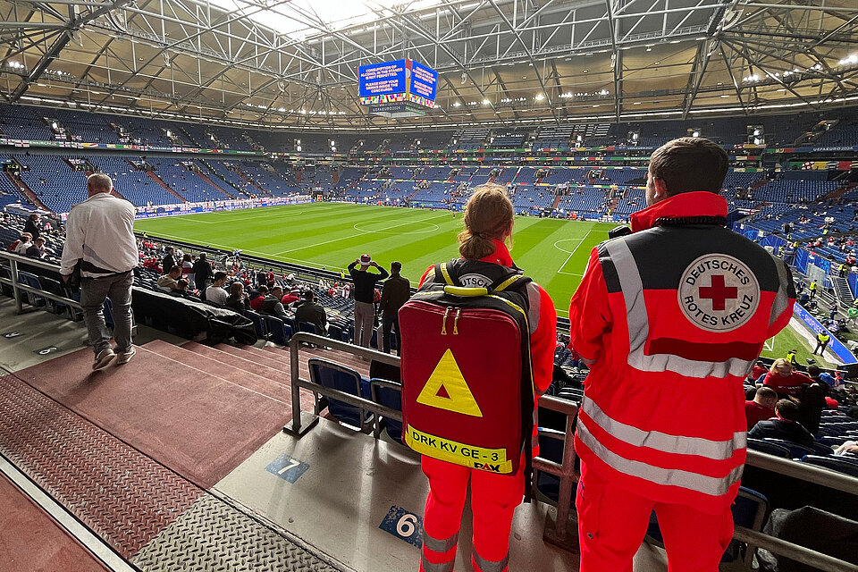 Zwei Mitglieder des Deutschen Roten Kreuzes stehen im Fußballstadion in Gelsenkirchen und blicken auf das Spielfeld. Sie tragen orangefarbene Jacken mit dem Logo des DRK auf dem Rücken, und einer von ihnen hat einen großen Notfallrucksack auf dem Rücken. Im Hintergrund sind einige Zuschauer zu sehen, und auf einer Anzeigetafel steht "UEFA EURO 2024".
