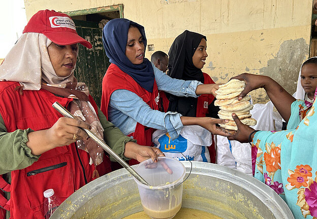 Drei Frauen vom Sudanesischen Roten Halbmond, erkennbar an ihren roten Westen, geben Essen aus. Eine Frau schöpft Suppe aus einem großen Metalltopf in einen Plastikbehälter, während die anderen Frauen einen Stapel Fladenbrot an eine Person in farbenfroher Kleidung überreichen. Im Hintergrund steht ein Kind in einem weißen Gewand.