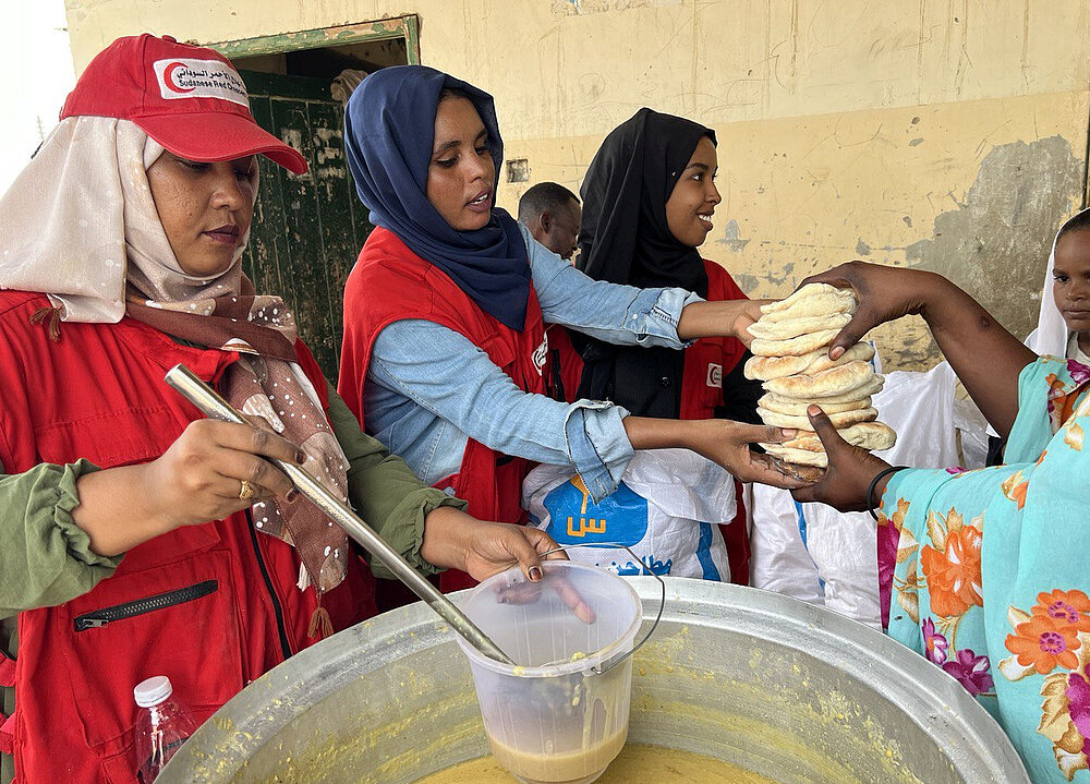 Drei Frauen vom Sudanesischen Roten Halbmond, erkennbar an ihren roten Westen, geben Essen aus. Eine Frau schöpft Suppe aus einem großen Metalltopf in einen Plastikbehälter, während die anderen Frauen einen Stapel Fladenbrot an eine Person in farbenfroher Kleidung überreichen. Im Hintergrund steht ein Kind in einem weißen Gewand.