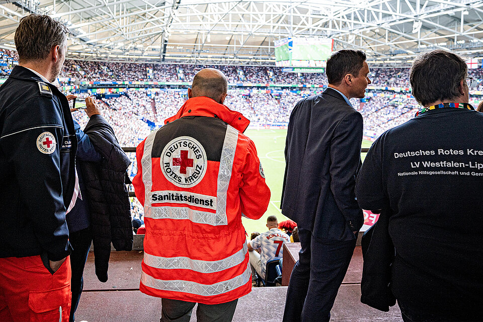 Mitglieder des Deutschen Roten Kreuzes stehen in einem Stadion und blicken auf das Spielfeld. Sie tragen verschiedene DRK-Uniformen, und das Stadion ist gefüllt mit Zuschauern.