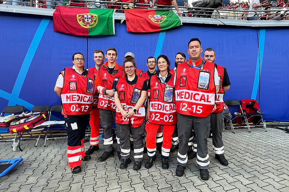 Ein Team von medizinischem Personal in roten Westen mit der Aufschrift "MEDICAL" und individuellen Nummern steht zusammen vor einer blauen Wand. Über ihnen hängen zwei portugiesische Flaggen. Im Hintergrund sind weitere Menschen zu sehen, die sich in einer Arena befinden.