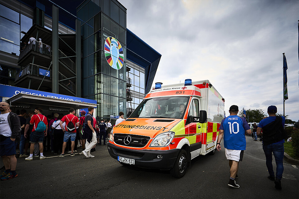 Ein Rettungswagen des Deutschen Roten Kreuzes steht vor der Arena auf Schalke. An der Wand des Stadions sieht man ein Logo der UEFA Euro 2024. Menschen in verschiedenen Trikots und Fanschals stehen in der Nähe des Fahrzeugs.