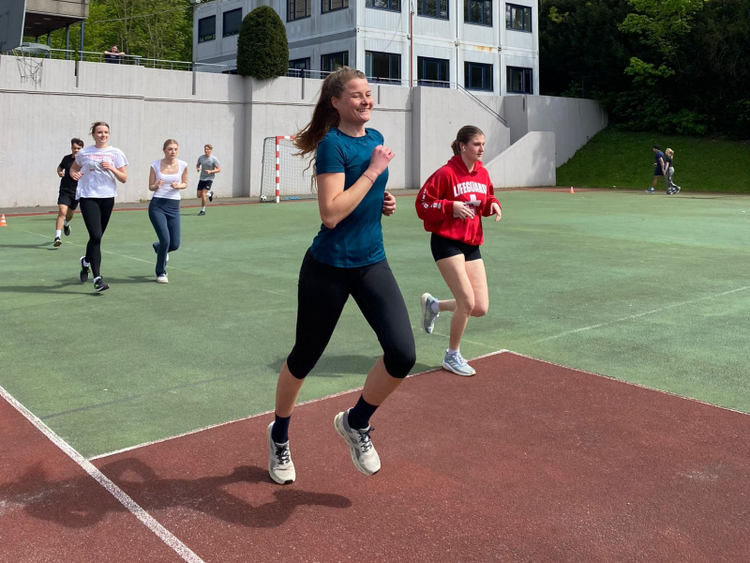 Das Bild zeigt einige Schülerinnen und Schüler, die anlässlich des Spendenlaufs der internationalen deutschen Schule in Brüssel um einen Sportplatz im Freien rennen. Die Sonne scheint, im Hintergrund sieht man hinter einer Mauer Schulgebäude. Die junge Läuferin im Vordergrund lächelt beim Laufen.