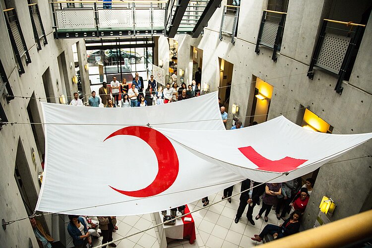 50 Jahrestag Rotes Kreuz Grundsätze im IFRK Büro Blick von oben in das Gebäude der Internationalen Föderation der Rotkreuz- und Rothalbmondgesellschaften. Über den Köpfen der versammelten Menschen hängen zwei große Stoffbahnen mit den Symbolen des Roten Kreuzes und des Roten Halbmonds.