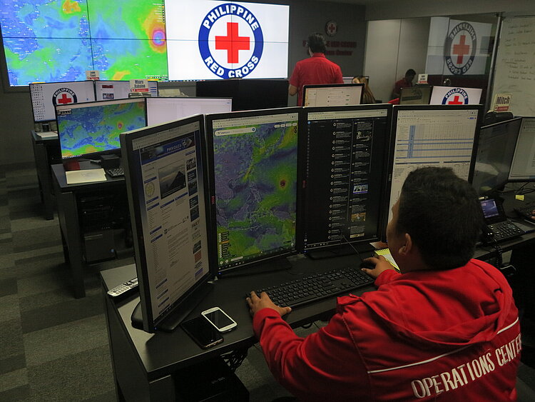 Ein Mitarbeiter des Philippinischen Roten Kreuzes arbeitet an mehreren Bildschirmen, auf denen Wetter- und Kommunikationsdaten angezeigt werden. Im Hintergrund sind weitere Mitarbeiter und ein großes Logo des Philippinischen Roten Kreuzes auf einem Bildschirm zu sehen.