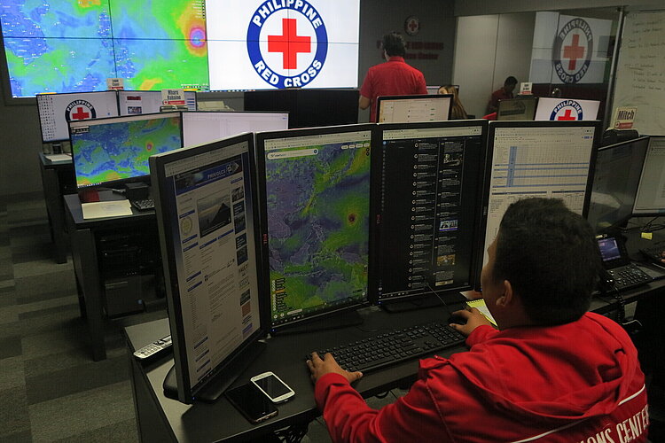 Einsatzkräfte des Philippinischen Roten Kreuzes Ein Mitarbeiter des Philippinischen Roten Kreuzes arbeitet an mehreren Bildschirmen, auf denen Wetter- und Kommunikationsdaten angezeigt werden. Im Hintergrund sind weitere Mitarbeiter und ein großes Logo des Philippinischen Roten Kreuzes auf einem Bildschirm zu sehen.