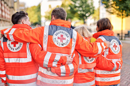 Vier Personen stehen in einer Reihe mit dem Rücken zur Kamera. Sie tragen rote DRK-Jacken und umfassen mit ihren Armen, die Person neben sich.