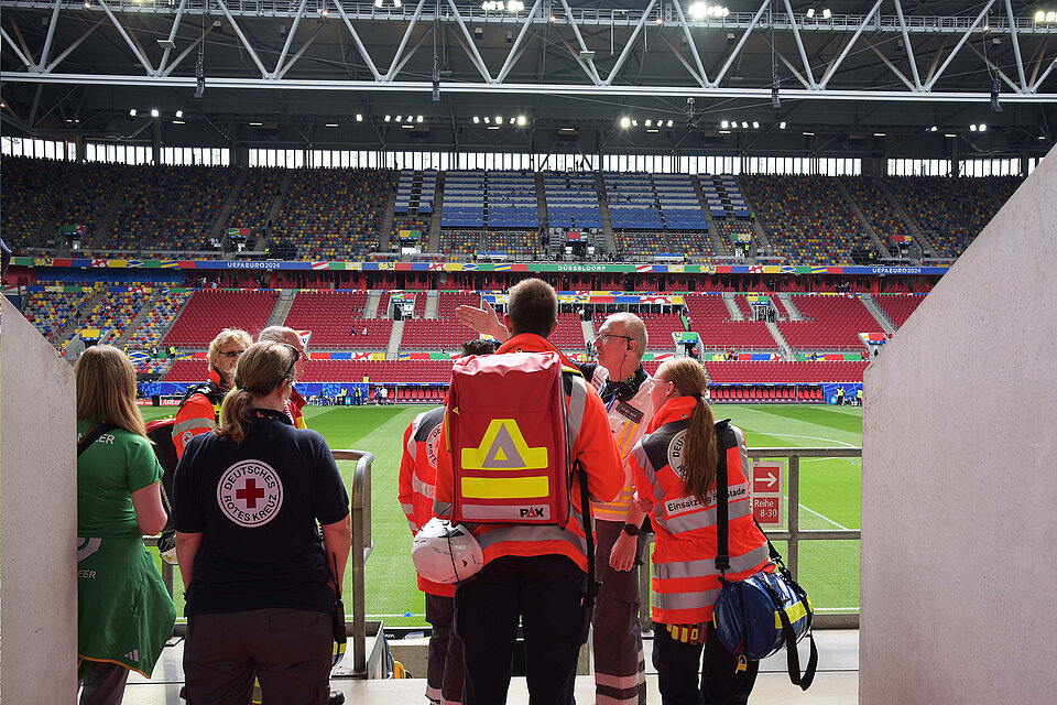 Rettungskräfte des Deutschen Roten Kreuzes stehen in einem Stadiontunnel und besprechen sich. Das Stadion im Hintergrund ist noch nicht gefüllt. Man sieht die bunten Sitzplätze. 