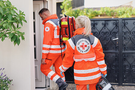 Zwei DRK-Rettungssanitäter betreten mit Notfallmaterialien ein Haus.