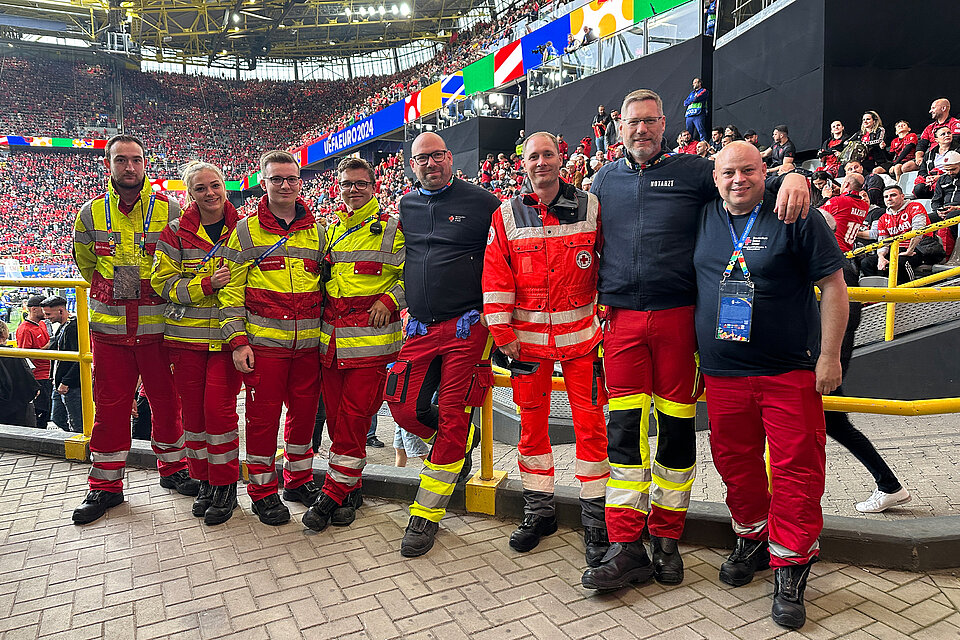 Eine Gruppe von acht Rettungskräften in roten und gelben Uniformen steht zusammen in einem vollen Stadion. Hinter ihnen sind zahlreiche Zuschauer und ein Bildschirm mit dem Schriftzug "UEFA EURO 2024" zu sehen. 