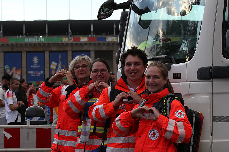 Vier Mitglieder des Deutschen Roten Kreuzes stehen nebeneinander vor einem Einsatzfahrzeug und formen mit ihren Händen Herzsymbole. Sie tragen orangefarbene Rettungsjacken und lächeln in die Kamera. Im Hintergrund sieht man das Olympiastadion in Berlin
