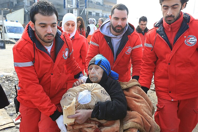 Mehrere Helfer vom Syrischen Arabischen Roten Halbmond in roter Kleidung evakuieren eine alte Frau