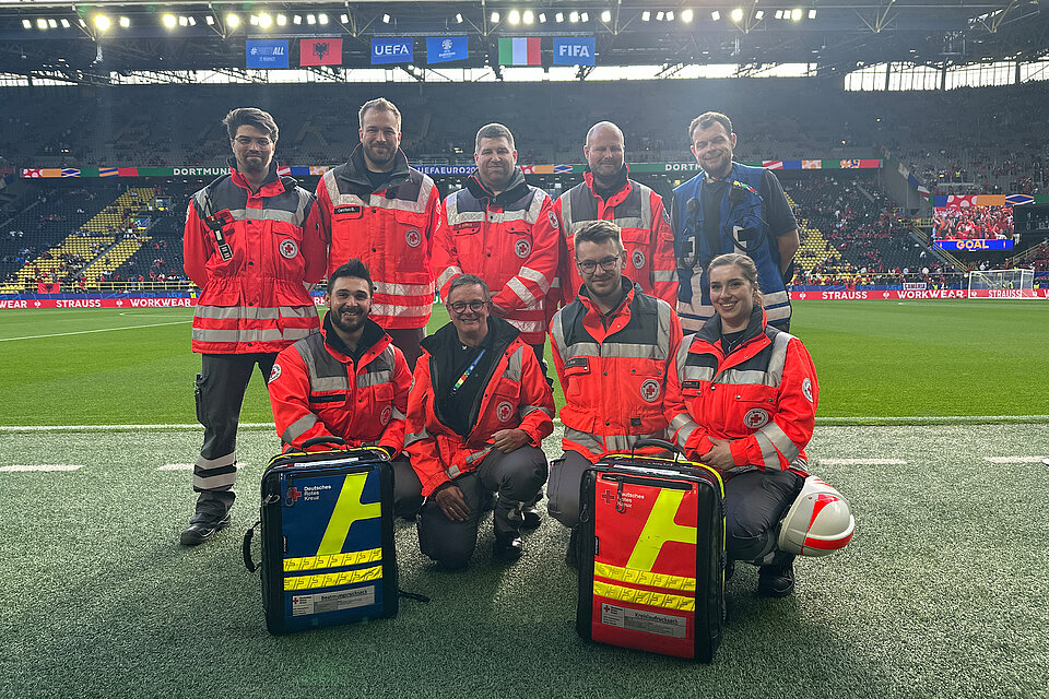 Eine Gruppe von neun Rettungskräften des Deutschen Roten Kreuzes posiert am Spielfeldrand eines Stadions. Sie tragen orangefarbene Jacken und lächeln in die Kamera. Vor ihnen liegen zwei große Notfallrucksäcke. Im Hintergrund sind das Spielfeld und die Tribünen mit Zuschauern zu sehen, sowie Anzeigetafeln mit den Logos der UEFA und FIFA.