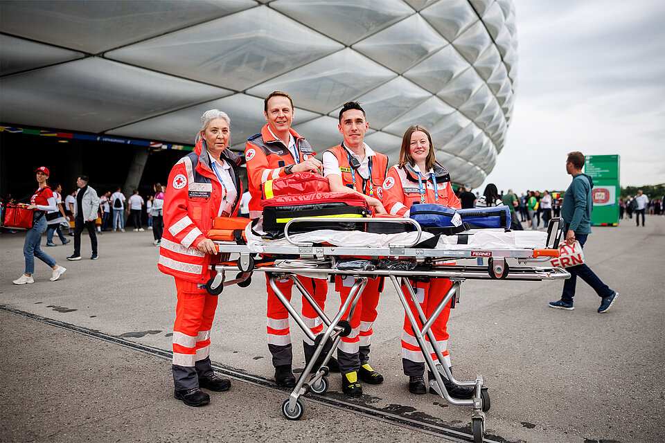 Vier Mitglieder des Deutschen Roten Kreuzes stehen vor der Allianz Arena in München und lächeln in die Kamera. Sie tragen orangefarbene Rettungsuniformen und stehen um eine Trage, die mit Notfallausrüstung beladen ist. 