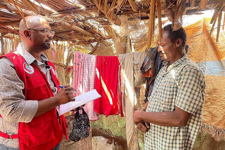 Ein Helfer des Sudanesischen Roten Halbmonds in roter Weste führt ein Gespräch mit einem lokalen Mann in einem karierten Hemd, umgeben von aufgehängter Wäsche in einer einfachen Unterkunft.