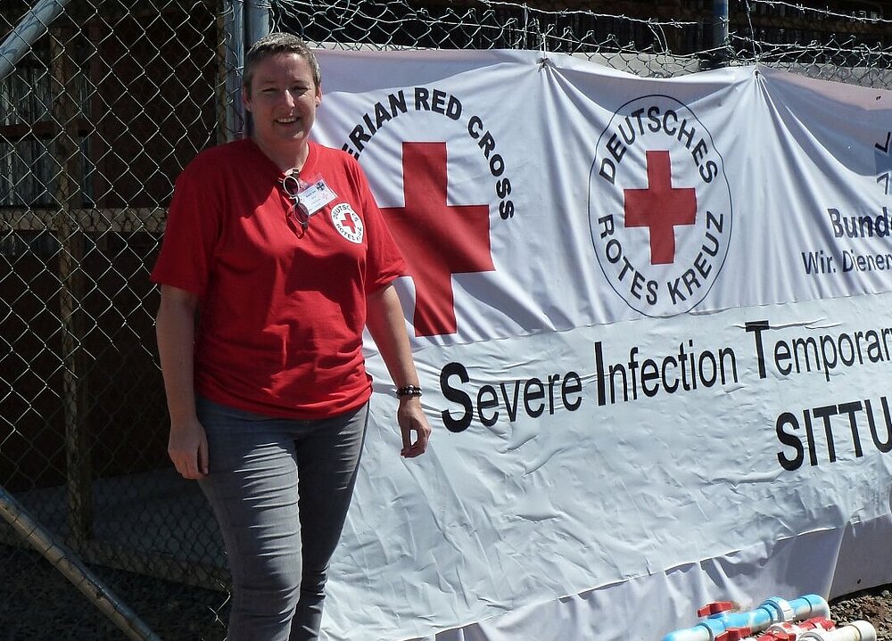 Eine Rotkreuz-Mitarbeiterin steht vor einem Zelt, auf dem die Logos des Liberianischen, des Deutrschen Roten Kreuzes und der Bundeswehr zu sehen sind.
