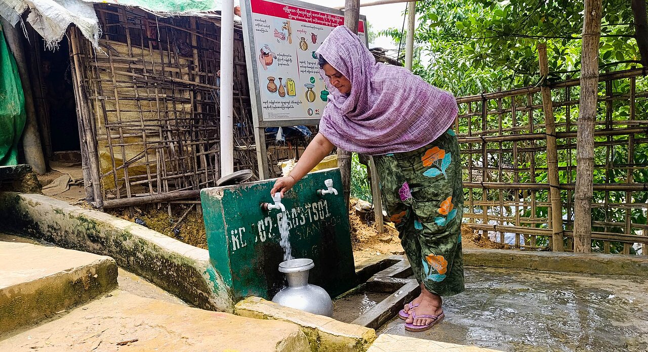 Wasserentnahmestelle_im_Fluechtlingscamp_Coxs_Bazar Eine Frau steht draußen an einer Wasserentnahmestelle und lässt Wasser in einen Blechkrug laufen